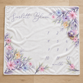 A square white baby blanket laid flat on a light wood floor, printed with the name 'Scarlett Blaise' in script at the top left, a floral border in pastel pink, purple, blue, and yellow around the edges, and a numbered monthly calendar from 1 to 12 with the word 'months' near the bottom.