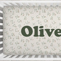 Custom baby crib bedding featuring an alphabet pattern with the name 'Olive' in large dark green letters on a cream background, shown inside a white wooden crib.