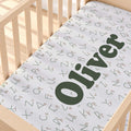 A personalized baby crib sheet with an all-over alphabet print and the name 'Oliver' in large dark green letters, shown fitted inside a light wood crib.