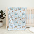 A personalized baby blanket with a repeating pattern of blue airplanes, brown mountains, and the name 'COLTON Joseph' printed in brown, draped over a light wood crib in a nursery setting.