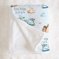 A personalized baby blanket with a white reverse side and a light blue printed front featuring watercolor-style airplanes, clouds, mountains, and the names 'COLTON joseph' in brown and blue lettering, laid flat on a white faux fur surface.