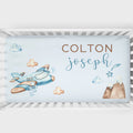 Personalized baby bedding set in a white crib, featuring a light blue background with watercolor illustrations of a blue airplane, clouds, stars, and a mountain. The text 'COLTON' is printed in brown block letters, and 'joseph' is written in blue cursive script.