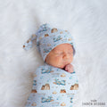 A sleeping newborn baby swaddled and wrapped in a light blue fabric with a printed pattern of mountains, clouds, birds, and the name 'COLTON joseph', wearing a matching tie-on hat. The baby is lying on a white faux fur blanket.
