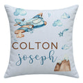 A square personalized throw pillow in light blue, featuring a watercolor-style blue and brown airplane, clouds, stars, and mountains. The text 'COLTON' is printed in brown block letters above 'joseph' in blue cursive script.