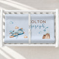 A white changing table with a personalized changing pad cover featuring a light blue background with a watercolor airplane, clouds, mountain, and custom names 'COLTON Joseph' in brown and blue lettering.