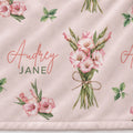 A close-up of a light blush pink fabric with a repeating floral print of pink gladiolus flowers and green leaves, plus the personalized name 'Audrey JANE' in coral script and green block letters.