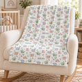 A personalized baby blanket with a colorful dinosaur and campfire pattern and the name 'adam' printed repeatedly, draped over a cream-colored rocking chair in a cozy nursery setting.
