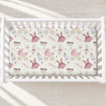 A baby crib with a fitted sheet featuring a personalized pattern of pink ballerina dresses, strollers, bottles, and bicycles, with the names 'Olivia' and 'Grace' printed repeatedly. The crib is shown from a top-down view on a light beige textured rug with soft sunlight shadows.