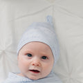A close-up of a baby lying on a white textured blanket, wearing a light blue knit hat with blue cursive text and a matching swaddle.