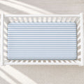 A top-down view of a white wooden crib with a light blue and white striped fitted sheet, set against a neutral cream textured rug with soft diagonal shadows.