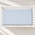 A light blue and white houndstooth patterned mattress protector fitted inside a white wooden baby crib, shown from a top-down perspective on a light textured rug.