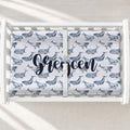 A personalized changing pad cover featuring a repeating blue whale print and the name 'Grecen' in large navy script across the center, shown from above in a bright nursery.