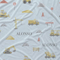 Light blue fabric with a construction-themed print featuring yellow dump trucks, excavators, cranes, traffic cones, and road signs, with the name 'ALONSO' printed in gray.