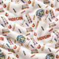 A close-up of a white fleece blanket with a school-themed pattern featuring books, apples, globes, crayons, scissors, paint palettes, and the name 'MORGAN' printed in red.
