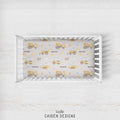A top-down view of a white wooden crib with a fitted sheet featuring a construction-themed print with yellow excavators, bulldozers, and the name 'MASON' repeated. The crib is set against a light, rustic white wood wall background.
