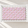 A pink baby crib mattress fitted sheet with a repeating pattern of chocolate chip cookies, milk bottles, and the name 'Emma' in brown, shown inside a white wooden crib.