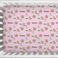 A pink crib fitted sheet with a repeating pattern of chocolate chip cookies, milk bottles, and the name 'Emma' in brown text, shown inside a white wooden crib.