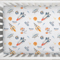 A fitted crib sheet with a colorful space-themed print featuring rockets, UFOs, planets, stars, and the personalized name 'Rowan' in blue script, shown inside a white wooden crib.