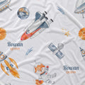 White fabric printed with a playful space-themed pattern featuring watercolor rockets, satellites, stars, planets, and a cartoon fox astronaut. The names 'Rowan QUINN' are printed in blue and orange text in two places.