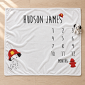 A personalized white baby blanket laid flat on a light wood floor, printed with the name 'HUDSON JAMES' at the top, a 12-month countdown grid, and firefighter-themed illustrations including a Dalmatian puppy in a red helmet and a red fire hydrant.