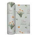 Pale sage swaddle blanket with white narcissus flowers, scattered green leaves, and the name Clara Noelle repeated in rose and green.