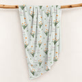 Pale sage swaddle blanket with white narcissus flowers, scattered green leaves, and the name Clara Noelle repeated in rose and green.