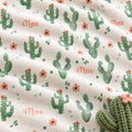 A close-up of a soft fleece blanket with a cactus pattern, featuring green pincushion cacti, small pink flowers, and the word 'Mae' in peach script. A knitted green cactus with a pink flower is visible in the bottom right corner.