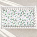 A white wooden crib with a fitted sheet featuring a green cactus and small orange flower pattern, with the name 'Mae' printed in orange script.