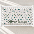A personalized baby crib mattress inside a white wooden crib, featuring a white background with a pattern of colorful alphabet letters and small dinosaur illustrations, with the name 'Oliver James' printed in dark green at the bottom.