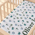 A personalized baby crib sheet with a white background, printed with colorful alphabet letters and dinosaur illustrations, shown fitted inside a light wood crib.