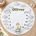 A round white baby milestone rug with a personalized name 'Oliver', numbers 1 through 12, watercolor clouds, a small green dinosaur, and the word 'months', shown laid out on a light wood floor.