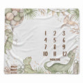 A square baby blanket with a white background featuring watercolor-style dinosaur illustrations, balloons, palm leaves, bunting, a birthday cake, and a printed monthly countdown from 1 to 12 labeled 'MONTHS'.