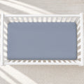 A top-down view of a light blue fitted crib mattress inside a white wooden crib, set on a textured cream rug with soft diagonal sunlight shadows.