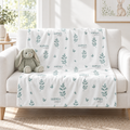 A white throw blanket with a repeating pattern of sage green leafy stems and the name 'SOPHIA printed in small dark text, draped over a light beige sofa in a bright living room. A plush bunny toy sits on the sofa.