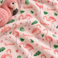 A close-up of a pink plush blanket or throw featuring a repeating flamingo print, green leaves, small pink flowers, and the word 'Leonor' in coral script. A matching pink plush flamingo toy is partially visible in the top left corner.
