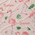 A close-up of a fabric surface featuring a repeating watercolor-style flamingo print on a light blush pink background, with green tropical leaves and coral script text reading 'Leonor'.