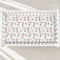 A white crib mattress fitted sheet with a repeating pattern of brown giraffes, green leaves, and the name 'ADAM' in brown text, shown inside a white wooden crib on a light textured floor.