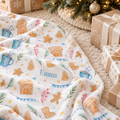A personalized white fleece blanket with a Christmas gingerbread print is draped over a beige carpet next to wrapped Christmas presents and a lit tree branch. The blanket features the name 'Emma' in blue script.