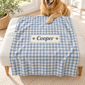 A blue and white gingham pet blanket with a custom name patch reading 'Cooper' is draped over a white ottoman in a bright room, with a golden retriever lying on the ottoman.
