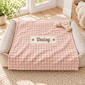 A personalized pet bed with a pink and white gingham dog blanket featuring a custom name patch that reads 'Daisy' with paw prints, set in a bright, sunlit room on a textured rug.