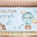 Personalized baby bedding set in a wooden crib, featuring a light blue duvet cover with watercolor illustrations of a hot air balloon, clouds, mountains, and stars, along with the names 'COLTON Joseph' in brown and blue text.