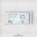 A personalized baby bedding set displayed in a white wooden crib, featuring a printed bassinet cover with watercolor illustrations of a hot air balloon, clouds, mountains, and the names 'COLTON joseph'.