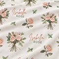A close-up of a cream-colored fabric with a repeating pattern of pink rose bouquets, green leaves, and the personalized text 'Isabella ROSE' in cursive and block lettering.