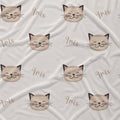 A close-up of a fabric surface with a repeating pattern of smiling cat faces and the word 'Iris' in cursive. The fabric is a warm off-white/cream color, and the cat faces are beige with dark pink/black ears, noses, and whiskers.