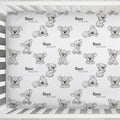 A personalized baby crib sheet with a repeating pattern of cute gray koala illustrations and names like 'Knox William', shown inside a white wooden crib.