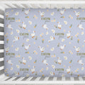 A fitted crib mattress cover with a repeating pattern of white geese, yellow flowers, and the text 'EVELYN grace' on a dusty lavender-blue background, shown inside a white wooden crib.