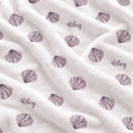A close-up of a soft white plush blanket with a repeating pattern of purple seashells and the name 'Riley' printed in script.