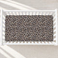 A leopard print crib mattress fitted inside a white wooden crib, shown from a top-down view on a light textured rug with soft window light shadows.