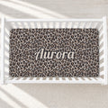 A top-down view of a white wooden crib containing a leopard print crib sheet with the name 'Aurora' personalized in the center. The background shows a light-colored floor with soft diagonal shadows.