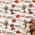 Western-themed fabric print featuring cacti, cowboy boots, cowboy hats, horseshoes, and red bandanas on a cream background, with the name 'WYATT' repeated. A plush brown horse toy is partially visible in the bottom right corner.